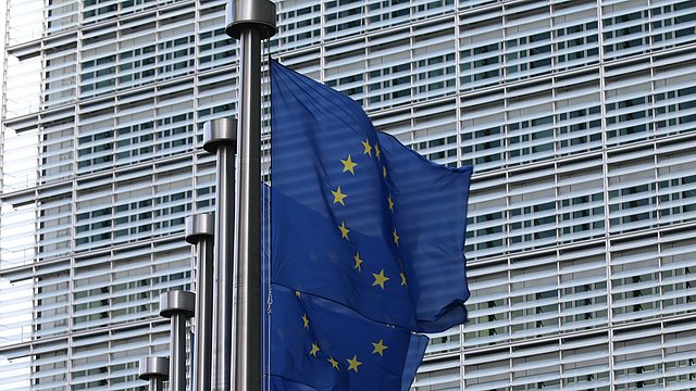 © Unsplash/Guillaume Périgois Das Bild zeigt eine Reihe von europäischen Flaggen mit einem Teil des Barlaymont-Gebäudes im Hintergrund
