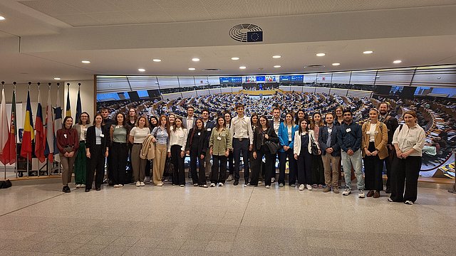 The image shows a group of students posing for a photo with an image of the hemicycle of the European Parliament in Brussels and the flags in the background.