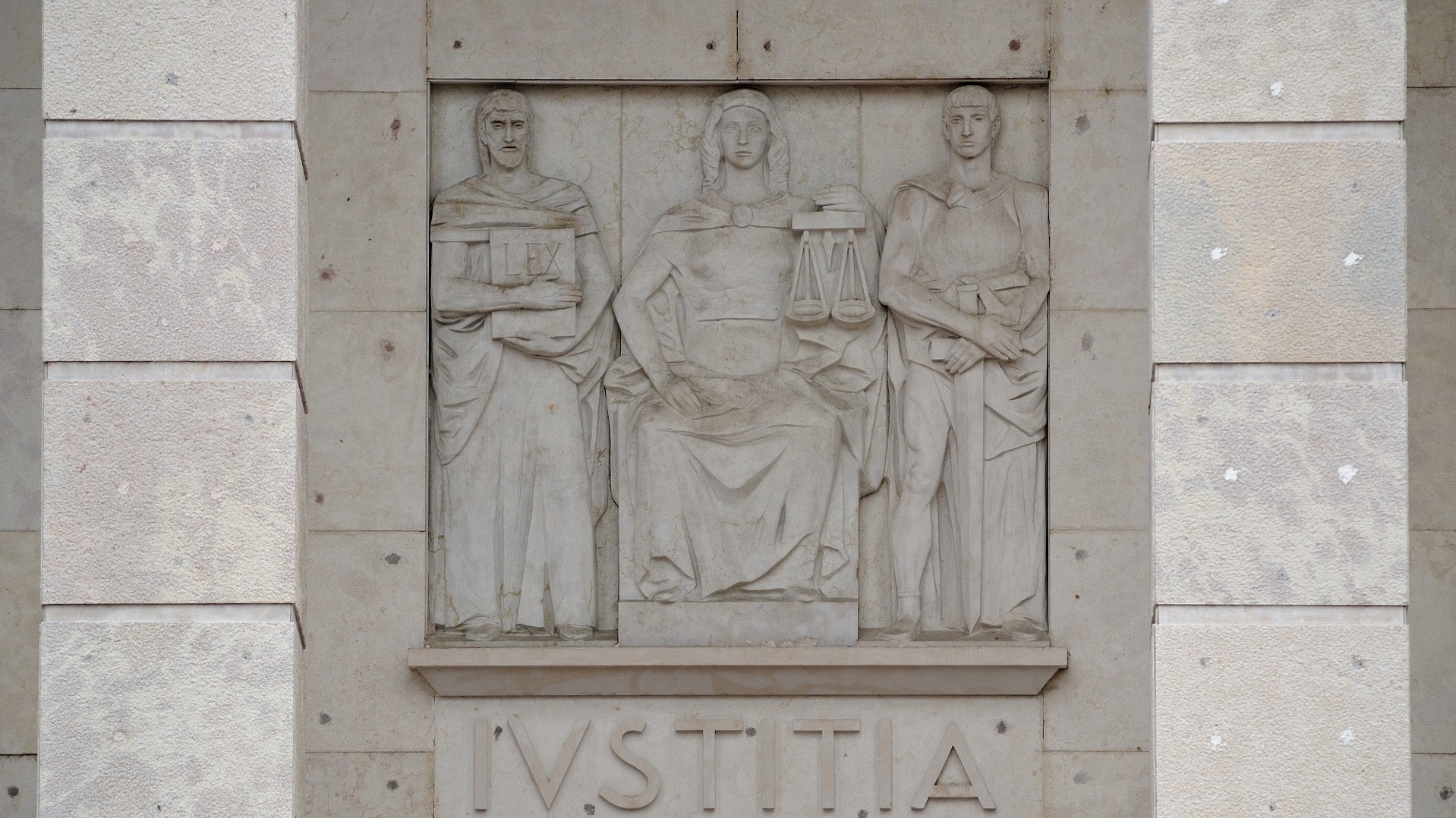Die sitzende Iustitia (Gerechtigkeit) am Gebäude des Bozner Landesgerichts, das ab 1939 errichtet wurde. Dieser fehlt auffälligerweise die Augenbinde, das Attribut, das für die Unparteilichkeit der Justiz steht.