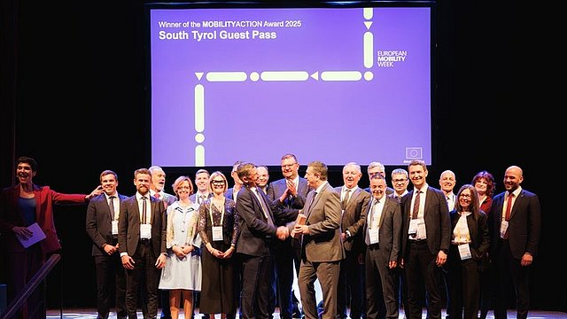 © LPA/Ivan Brentgani La delegazione altoatesina è sul palco e alcuni dei suoi membri applaudono. Al centro della delegazione c’è l’Assessore provinciale alla Mobilità Daniel Alfreider, che tiene in mano un trofeo. Dietro di lui c’è uno schermo su cui è scritto «Winner of the MOBILITYACTION Award 2025 South Tyrol Guest Pass».