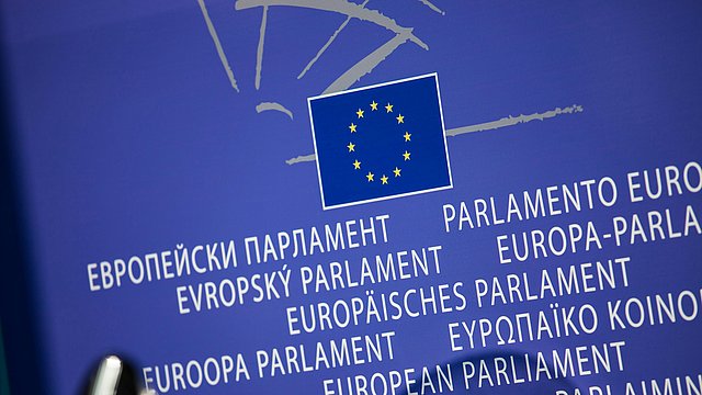 Das Bild zeigt einen blauen Hintergrund mit dem Logo der Europäischen Union in der Mitte. Unter dem Logo steht der Schriftzug „Europäisches Parlament“ in mehreren Amtssprachen der EU. Im Hintergrund ist eine Grafik zu sehen, die die Struktur des Europäischen Parlaments darstellt. 