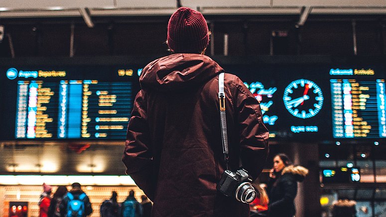 Junger Mann steht mit Rucksack vor einer Anzeigetafel am Bahnhof.