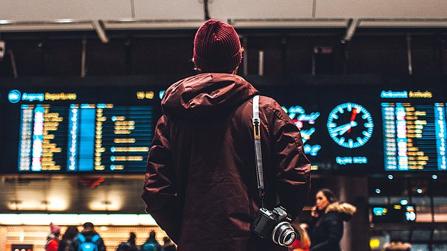 © Unsplash/Erik Odiin - Ragazzo di spalle con cappelli e macchina fotografica sulla spalla guarda il tabellone delle partenze della stazione dei treni.