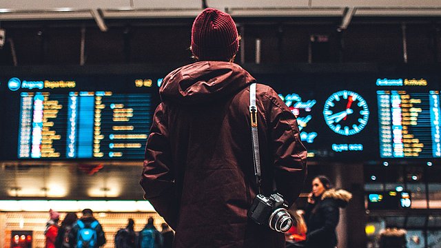 © Unsplash/Erik Odiin Junger Mann steht mit Rucksack vor einer Anzeigetafel am Bahnhof.