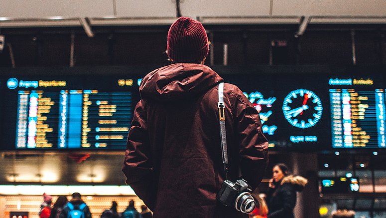 - Ragazzo di spalle con cappelli e macchina fotografica sulla spalla guarda il tabellone delle partenze della stazione dei treni.