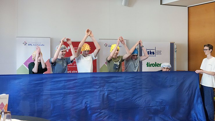 SchülerInnen mit Bademasken heben Hände hoch, davor blaues Tuch (steht für Wasser)