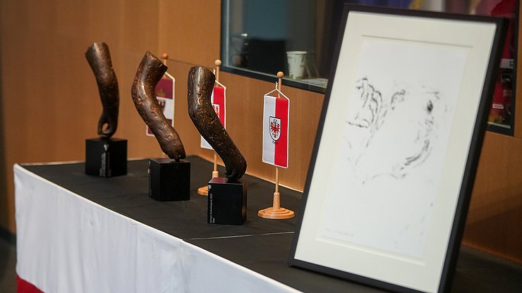 Im Bild die Preise für die Gewinner:innen des Euregio-Baukulturpreises: (von links) drei von Lois Anvidalfarei geschaffene Bronzeskulptur und der Publikumspreis und zwar eine Lithografie des ladinischen Künstlers Anvidalfarei.
