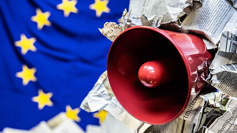Un megafono passa attraverso una pagina di giornale e la bandiera dell’UE sullo sfondo