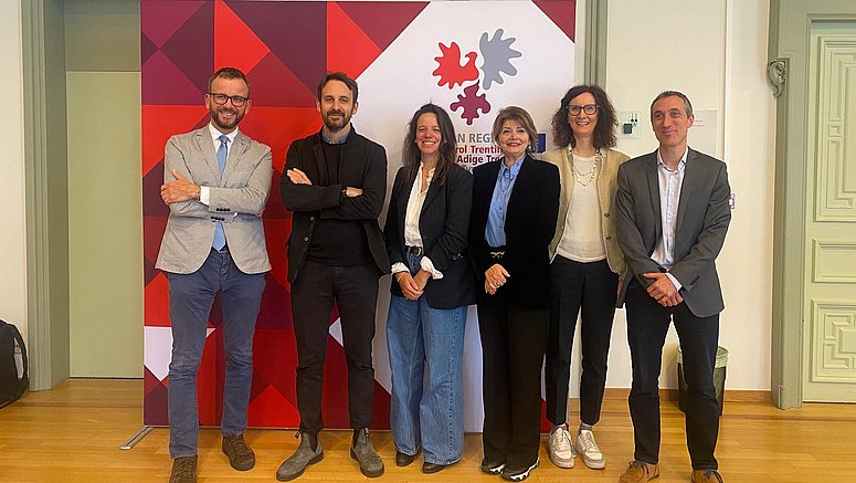 Das Foto zeigt einige der Redner*innen und Teilnehmer*innen der Veranstaltung in einer Reihe nebeneinander, vor dem Logo der Vertretung der Europaregion Tirol - Südtirol - Trentino.