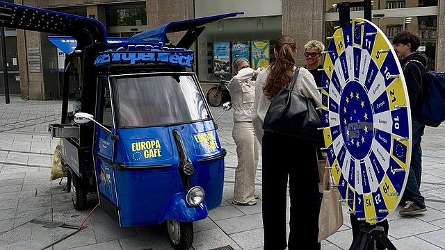 © Land Tirol Auf der rechten Seite des Bildes ist ein blau, gelb und weiß gefärbtes Glücksrad zu sehen; hinter dem Rad steht eine Gruppe von vier Personen, die sich unterhalten und lachen. Auf der linken Seite des Bildes ist ein blauer Apecar zu sehen, mit Flügeln über dem Dach und gelben Schriftzügen an der Vorderseite, auf denen „Europa Café“ steht.