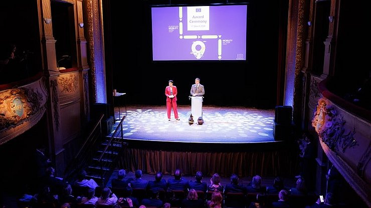 In der Bildmitte ist der EU-Kommissar für nachhaltigen Verkehr und Tourismus, Apostolos Tzitzikōstas, zu sehen, links von ihm die Moderatorin. Über dem EU-Kommissar befindet sich eine Leinwand, auf die die Präsentation projiziert wird. Im unteren Bildbereich sitzen die Zuschauer. Die Sitzreihen des Theaters befinden sich rechts und links im Bild.