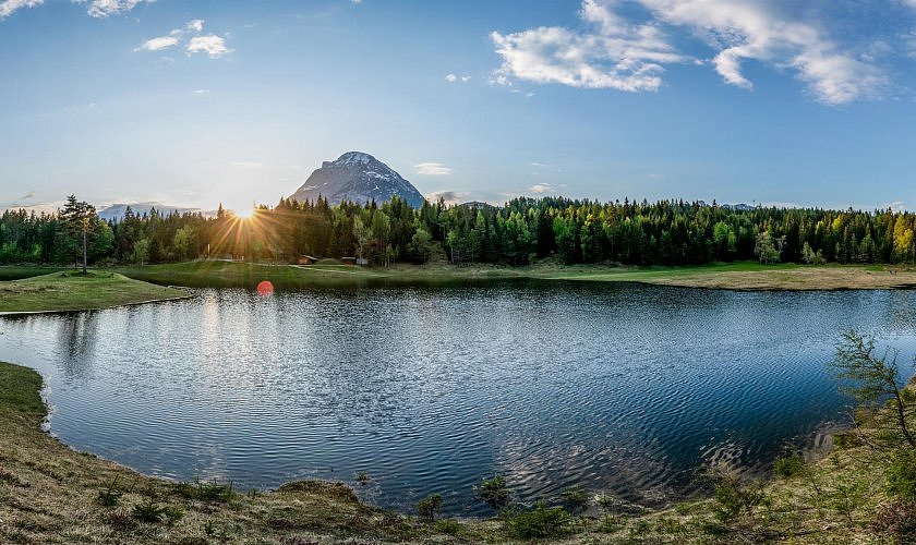 Ein Bergsee bei Sonnenuntergang mit Blick auf einen Wald und dahinter einen einzelnen Berg.  