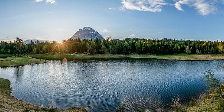 Ein Bergsee bei Sonnenuntergang mit Blick auf einen Wald und dahinter einen einzelnen Berg.  