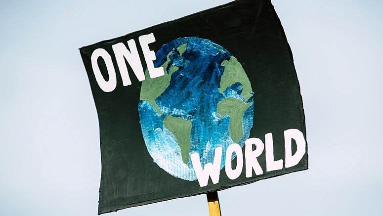 Un cartello di protesta dipinto a mano mostra un’illustrazione della Terra in blu e verde su uno sfondo scuro. Sopra e sotto l’immagine compaiono in grandi lettere bianche le parole “ONE WORLD”. Il cartello è sollevato con un bastone di legno contro il cielo chiaro. 