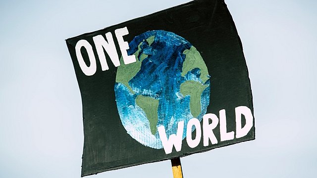 Un cartello di protesta dipinto a mano mostra un’illustrazione della Terra in blu e verde su uno sfondo scuro. Sopra e sotto l’immagine compaiono in grandi lettere bianche le parole “ONE WORLD”. Il cartello è sollevato con un bastone di legno contro il cielo chiaro. 