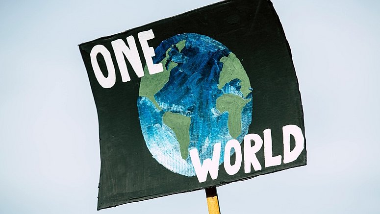 Ein handgemaltes Protestschild zeigt eine blau-grüne Darstellung der Erde auf dunklem Hintergrund. Darüber und darunter stehen in großen weißen Buchstaben die Worte „ONE WORLD“. Das Schild wird an einem Holzstab gegen den hellen Himmel hochgehalten. 