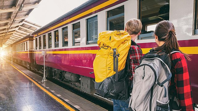 Un ragazzo e una ragazza di spalle aspettano sul binario davanti ad un treno rosso